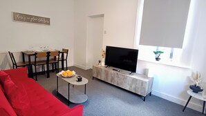 A modern living room with a red sofa, a dining table with chairs, a TV on a wooden stand, and a window with a plant. - Prime Town Centre-Location-Station-Airport-WiFi-Parking (Luton)