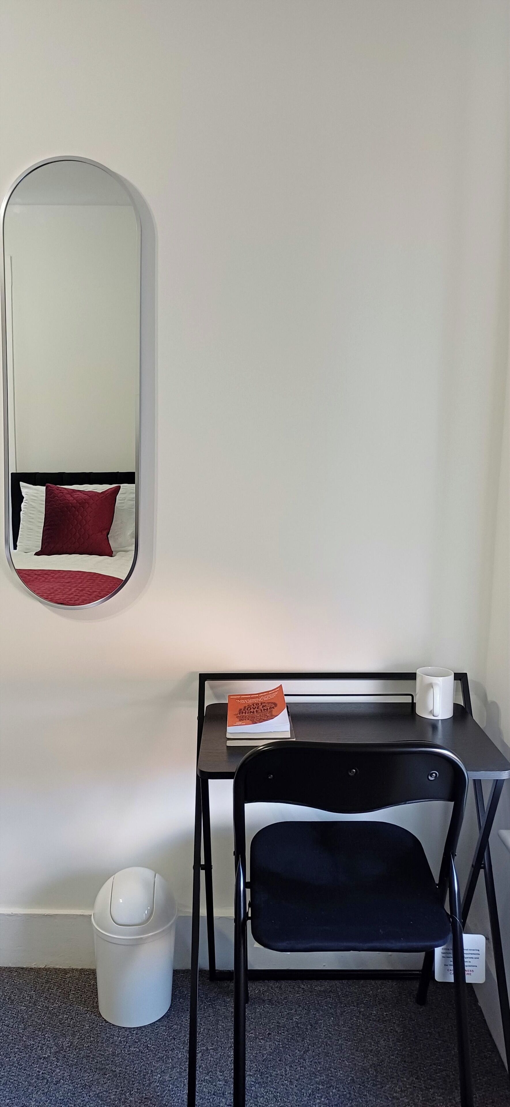 A compact room with a bed, a small desk with a chair, a white trash bin, and a wall-mounted mirror.