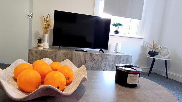 A living room with a TV on a stand, a bowl of oranges, and a candle on a table. - Prime Town Centre-Location-Station-Airport-WiFi-Parking (Luton)