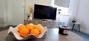 A living room with a TV on a stand, a bowl of oranges, and a candle on a table. - Prime Town Centre-Location-Station-Airport-WiFi-Parking (Luton)