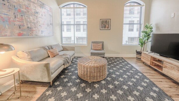 Ein Wohnzimmer mit Sofa, Hocker, Fernsehschrank und großen Fenstern.