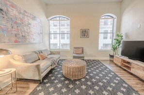 A living room with a sofa, ottoman, TV stand, and large windows. - Milk153-2C-2/2 · Airy 2BR | Fidi Boston | Walk anywhere! (Boston)