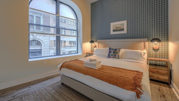 A bedroom with a large arched window, a bed with a brown throw blanket, two bedside lamps, and a tiled wall. - Bold, Spacious 2br in Boston`s Financial District (Boston)