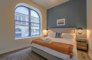 Une chambre avec une grande fenêtre en arc de cercle, un lit recouvert d’un plaid marron, deux lampes de chevet et un mur recouvert de carreaux.
