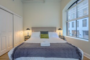 A neatly made bed with a headboard, two bedside lamps, a window with blinds, and a door. - Bold, Spacious 2br in Boston`s Financial District (Boston)