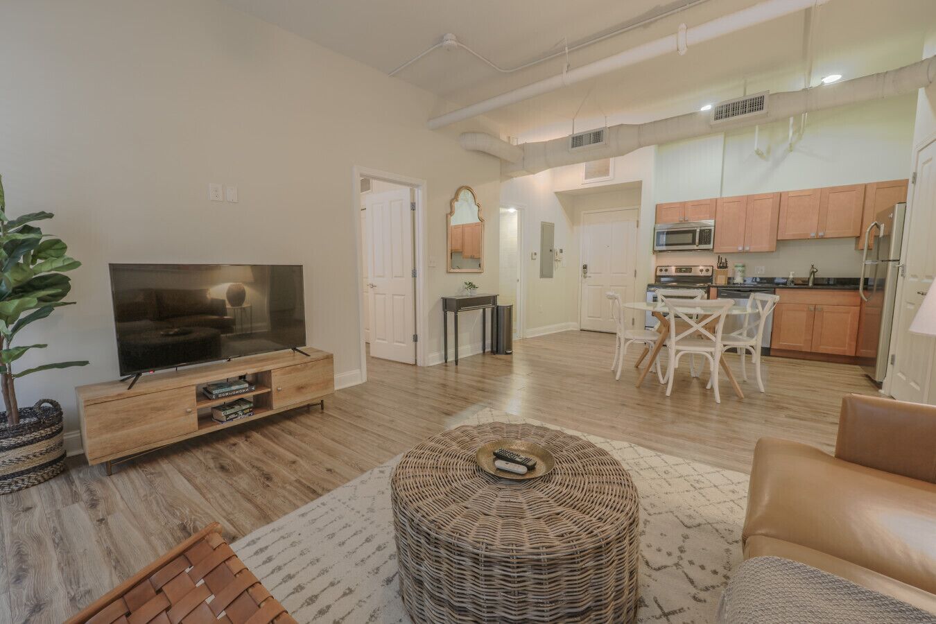 A modern living room with a flat-screen TV, a wooden entertainment unit, a wicker ottoman, and a brown leather sofa.
