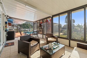 Uma área de varanda coberta com móveis de vime, uma mesa de vidro e vista para o quintal.