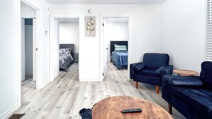 En moderne, åben stue med sofa, lænestol og sofabord. To soveværelser er synlige, hvoraf det ene har en seng og det andet et skrivebord og stol.