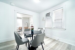 A modern dining area with a round table and six chairs, a vase with flowers, and a view of a living room with a sofa and a chair. - 1032 Assumption · Trendy 3 BedRoom Perfect for Long Stays (Windsor)