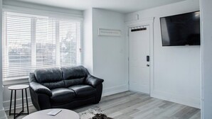 A living room with a black leather sofa, a small round table, a mounted TV, and a door with a coat rack. - 1036 Assumption · Bright 3 BDRM Unit Perfect for Longer Stays (Windsor)
