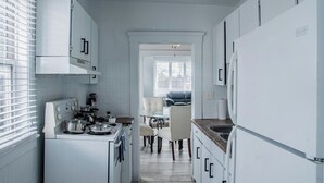 A narrow kitchen with white cabinets, a stove, and a dining area with a table and chairs. - 1036 Assumption · Bright 3 BDRM Unit Perfect for Longer Stays (Windsor)