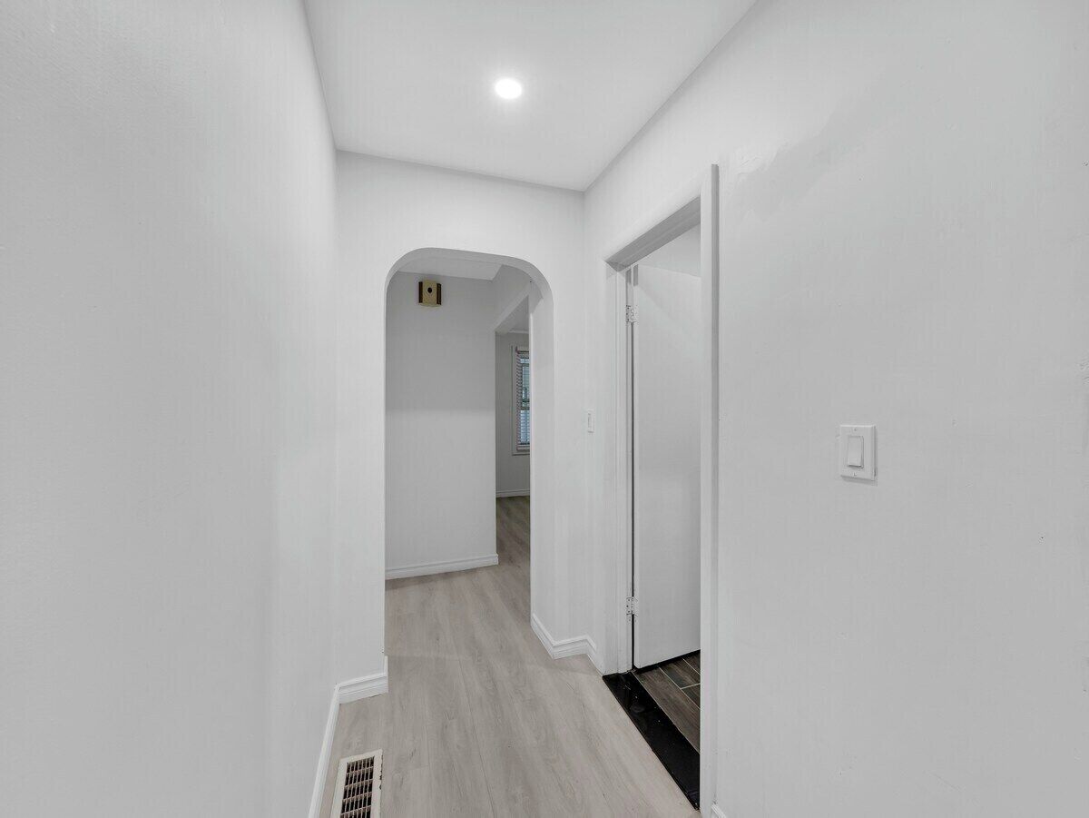Un couloir aux murs blancs et au plancher en bois, qui mène à une pièce dotée d’une porte.