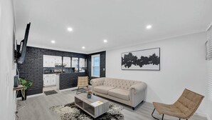 Uma sala de estar moderna com um sofá, mesa de centro e uma poltrona. Há uma área de cozinha com armários e bancada. Uma grande obra de arte urbana está na parede.