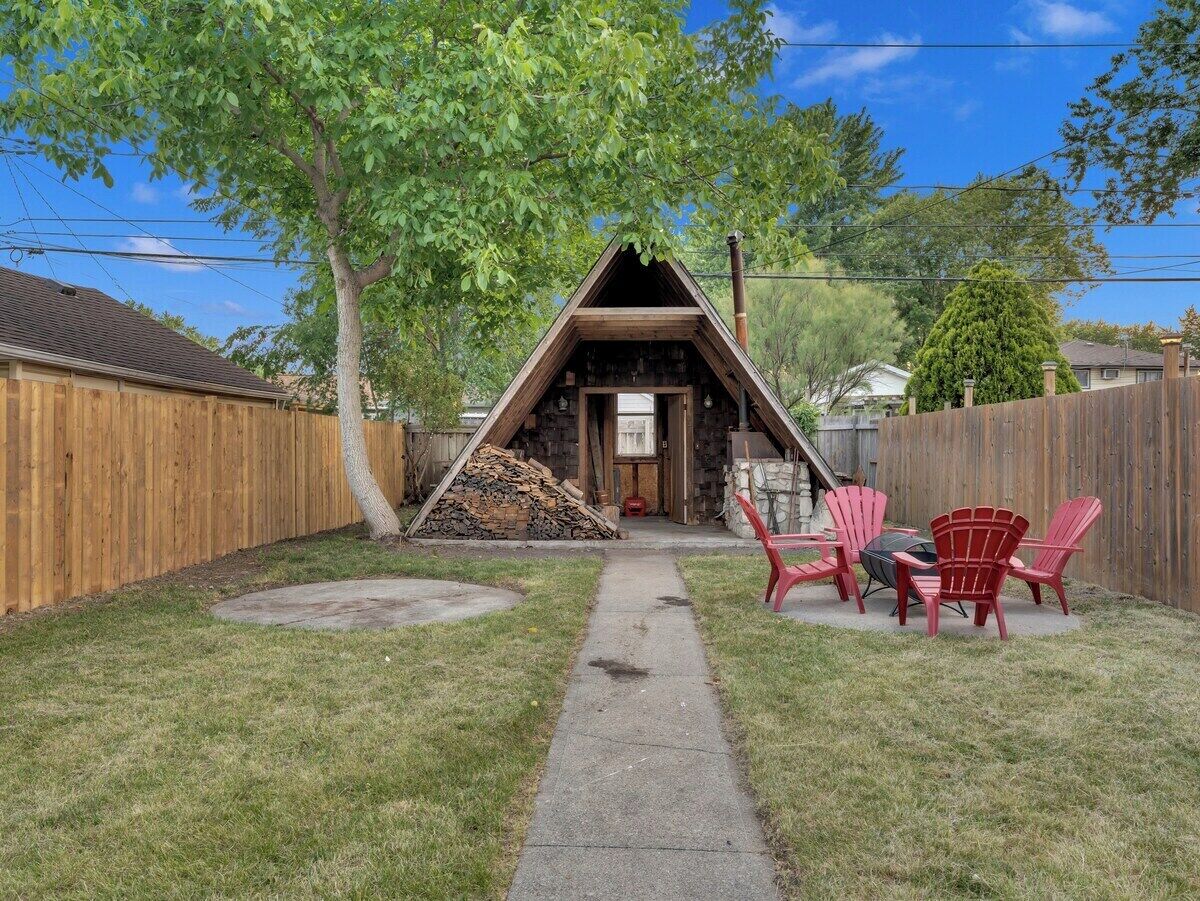 A triangular wooden structure with a chimney, a fire pit, and red Adirondack chairs in a backyard.