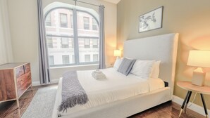 A modern bedroom with a large bed, a wooden dresser, a small table with a lamp, and a window with curtains. - Milk153-3D-2/2 · 2br/2ba FIDI Apt | Keyless Entry | Walk anywhere! (Boston)