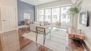 A modern living room with hardwood flooring, a grey sofa, a glass coffee table, and a large window with a city view. - Milk153-3D-2/2 · 2br/2ba FIDI Apt | Keyless Entry | Walk anywhere! (Boston)