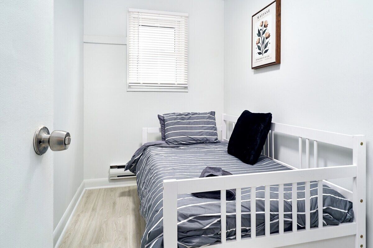 A small, neatly arranged bedroom with a single bed, a window with blinds, a framed picture on the wall, and a door with a silver handle.