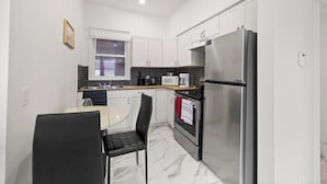A compact kitchen with white cabinets, a stainless steel refrigerator, a microwave, and a small dining area with a table and chairs. - 219 · New Build Cozy 2 Bedroom Unit (Windsor)