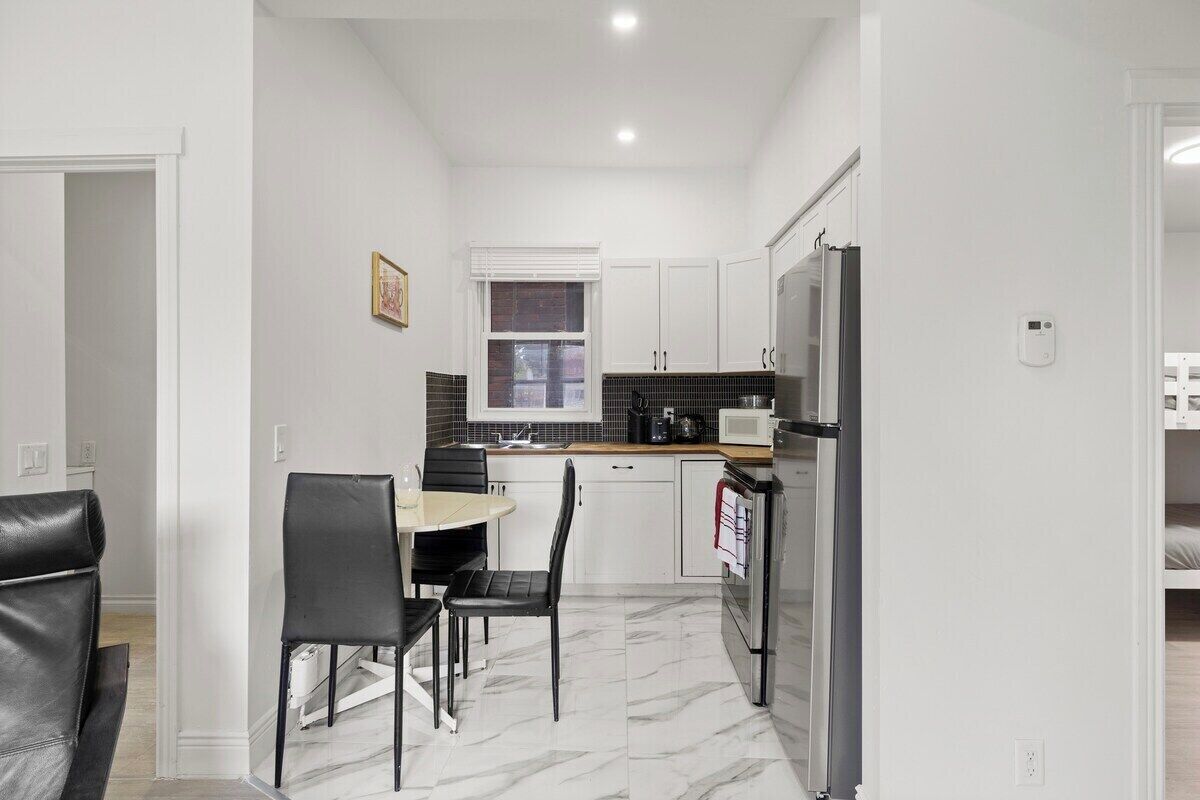 Cozinha compacta com armários brancos, bancada preta, geladeira e um espaço de refeições com mesa e cadeiras.