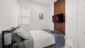 A modern bedroom with a bed, a TV mounted on the wall, a window with blinds, and a wooden panel wall. - 219 · New Build Cozy 2 Bedroom Unit (Windsor)