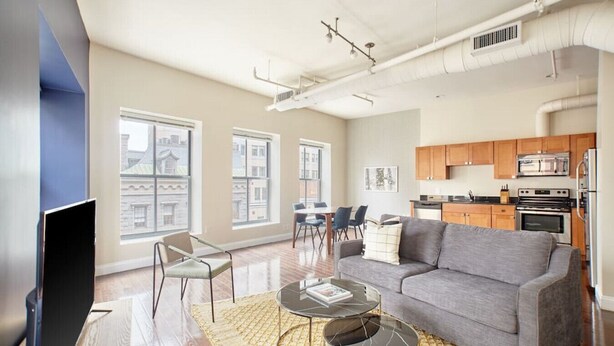 Una sala de estar moderna con un sofá gris, una mesa de centro y un televisor. Hay ventanas amplias, un comedor y una cocina con armarios de madera.