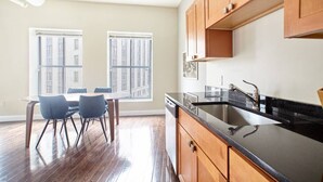Cocina moderna con armarios de madera, encimera negra y pileta de acero inoxidable. Cuenta con un comedor con mesa y sillas, y ventanas amplias que ofrecen luz natural.