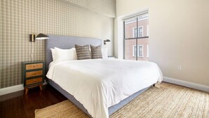 Un dormitorio con una cama grande, una mesita de noche, una lámpara y vistas a un edificio de ladrillo a través de una ventana.