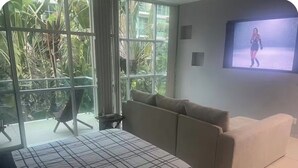 A bedroom with a bed, a sofa, a TV, and a window with a view of greenery.