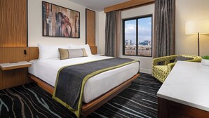 A hotel room with a bed, a desk, a chair, a window with a city view, and a painting on the wall.