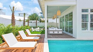 Zona de piscina exterior moderna amb gandules de fusta, una petita zona de seients amb coixins blancs i un porxo amb vidres.