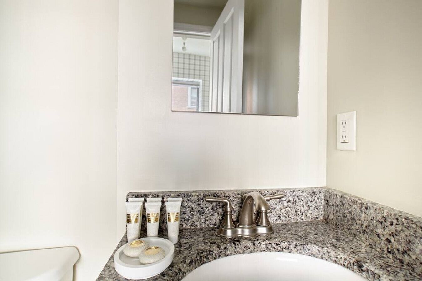 Une salle de bain avec un comptoir en granit, un lavabo blanc et un miroir au-dessus du lavabo. On y trouve trois flacons de lotion sur le comptoir.