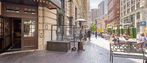 A street view with a building labeled "PUBLIC HOUSE" and a "BOSTONIA" sign. There are outdoor seating areas with tables and umbrellas, and people are visible in the distance.