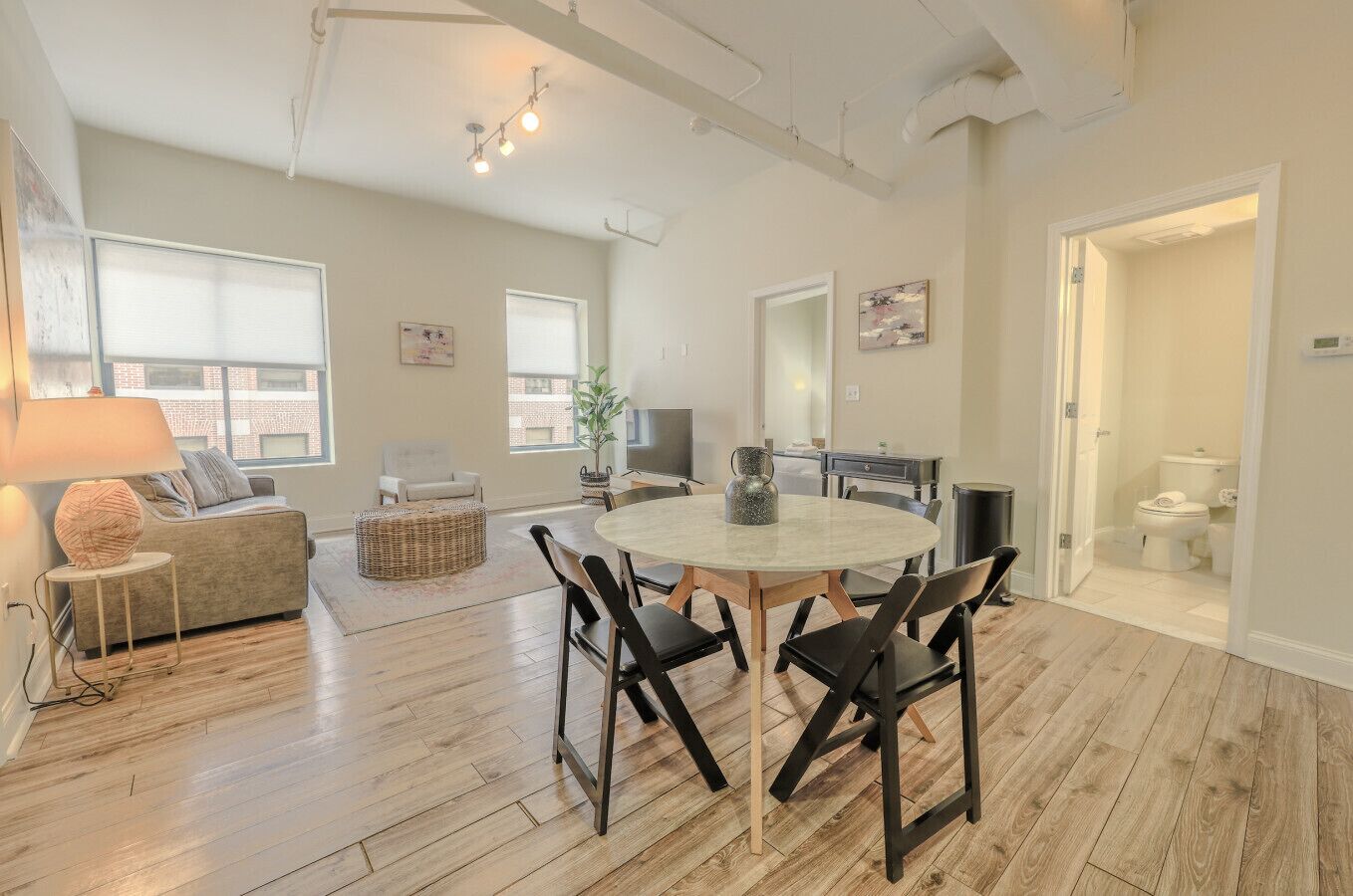 A spacious living area with a round dining table, black chairs, a sofa, and a coffee table. There is a TV, a plant, and a window providing natural light.