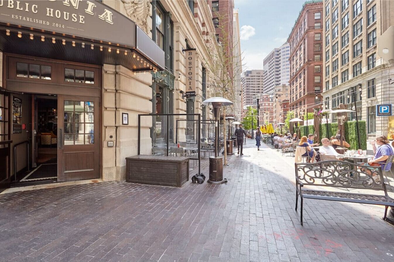 A street with outdoor seating, a building with a 'PUBLIC HOUSE' sign, and a 'BOSTONIA' sign on a pole.