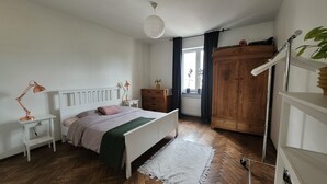 Quarto com cama, mesas de cabeceira, guarda-roupa, radiador e janela com cortinas.