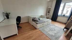 Quarto compacto com cama, escrivaninha, cadeira, uma pequena planta, estante e janela com cortinas.
