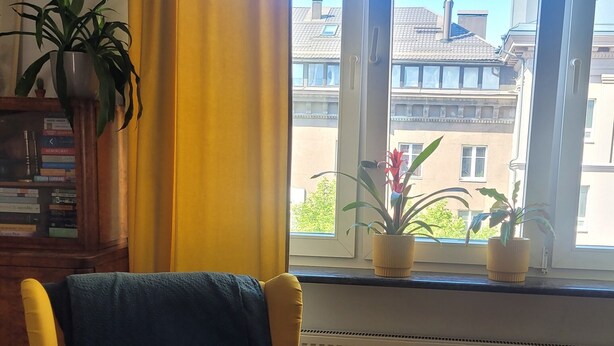 Uma poltrona amarela com almofada escura em um quarto com uma janela grande, um radiador e um vaso de planta no parapeito.