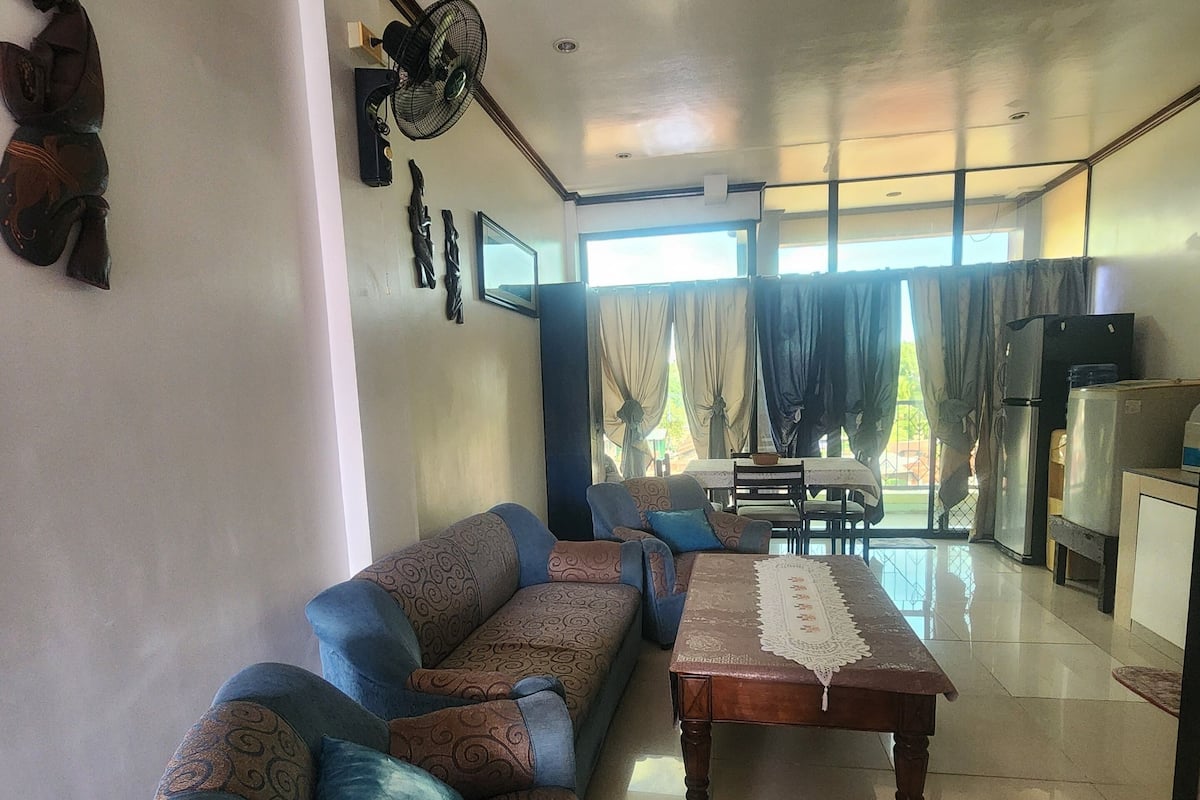 Una sala de estar con sofá, una mesa de centro y una mesa de comedor. Hay un ventilador de techo, un refrigerador y una ventana con cortinas.
