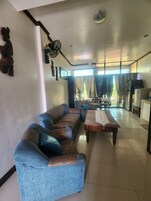Una sala de estar con sofá, una mesa de centro y una mesa de comedor. Hay un ventilador de techo, un refrigerador y una ventana con cortinas.