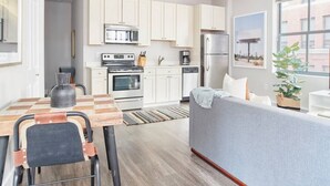 Une cuisine moderne avec des armoires blanches, un four encastré, un micro-ondes et un réfrigérateur. On y trouve un coin repas avec une table et des chaises en bois, ainsi qu’un canapé gris dans l’espace salon.