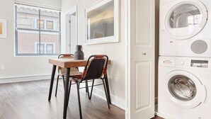 Une buanderie équipée d’un lave-linge et d’un sèche-linge superposés, d’une table en bois avec des chaises et de tableaux encadrés accrochés au mur.