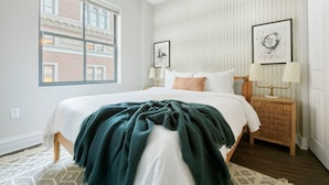 Une chambre avec un grand lit, deux tables de chevet avec des lampes, une fenêtre donnant sur un immeuble et un tapis à motifs.