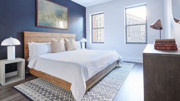 A modern bedroom with a large bed, a wooden headboard, a painting above the bed, and two windows with a view of buildings. - Milk153-6G-1/1 · Bright FIDI 1br | Centrally Located (Boston)