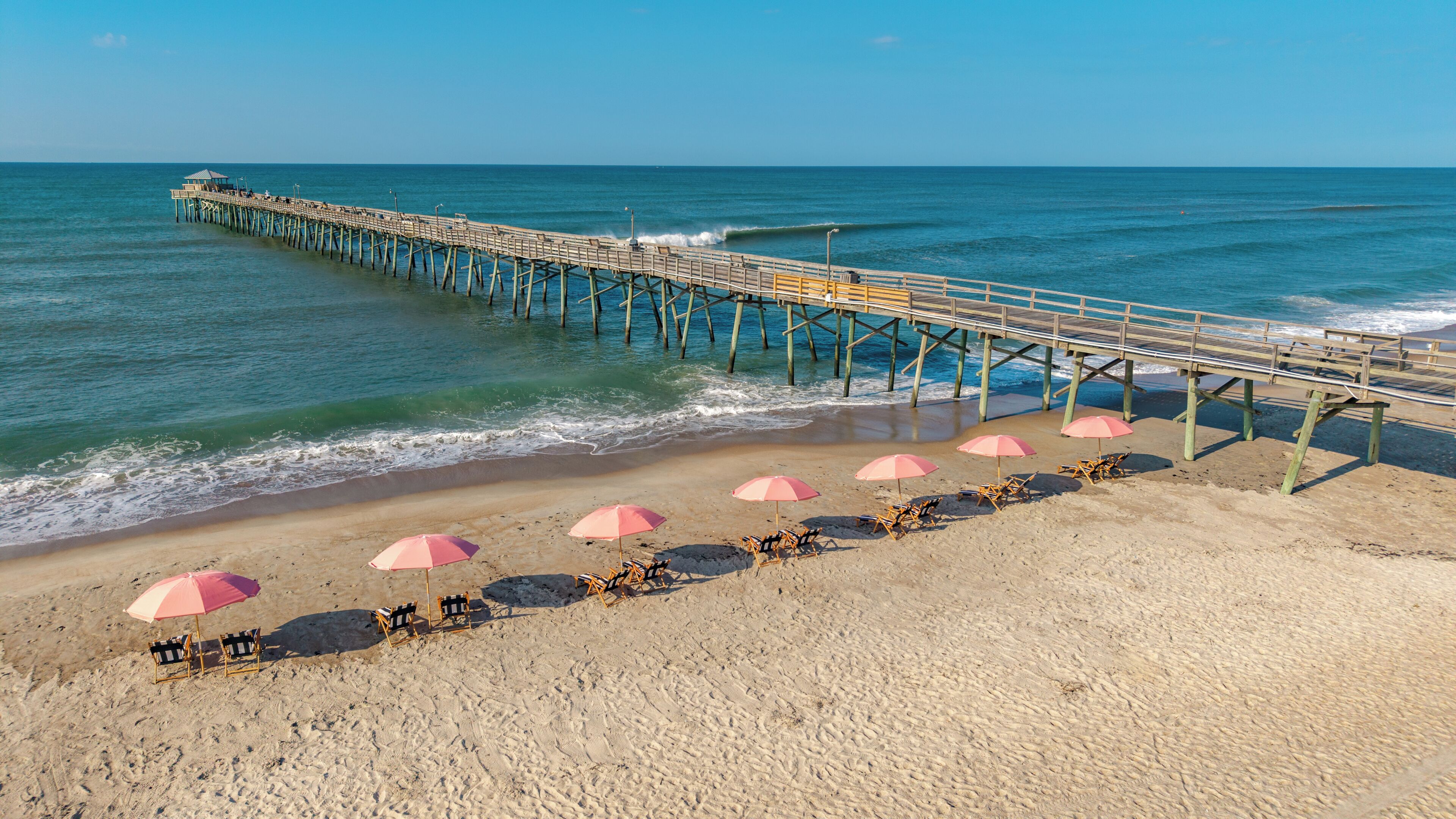 A beach with a wooden pier extending into the ocean, accompanied by beach chairs and umbrellas.