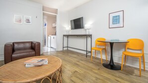 A room with a brown leather sofa, a wicker table with magazines, a small round table with two orange chairs, a flat-screen TV on a stand, and a framed picture on the wall. - Oceanana Motel (Atlantic Beach)