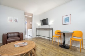 Ein Raum mit einem braunen Ledersofa, einem Korbtisch mit Zeitschriften, einem kleinen runden Tisch mit zwei orangefarbenen Stühlen, einem Flachbildfernseher auf einem Schrank und einem gerahmten Bild an der Wand.