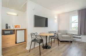 Ein kleines Zimmer mit Küchenzeile, Flachbildfernseher, zwei Stühlen, einem kleinen Tisch und einem Fenster mit Vorhängen.