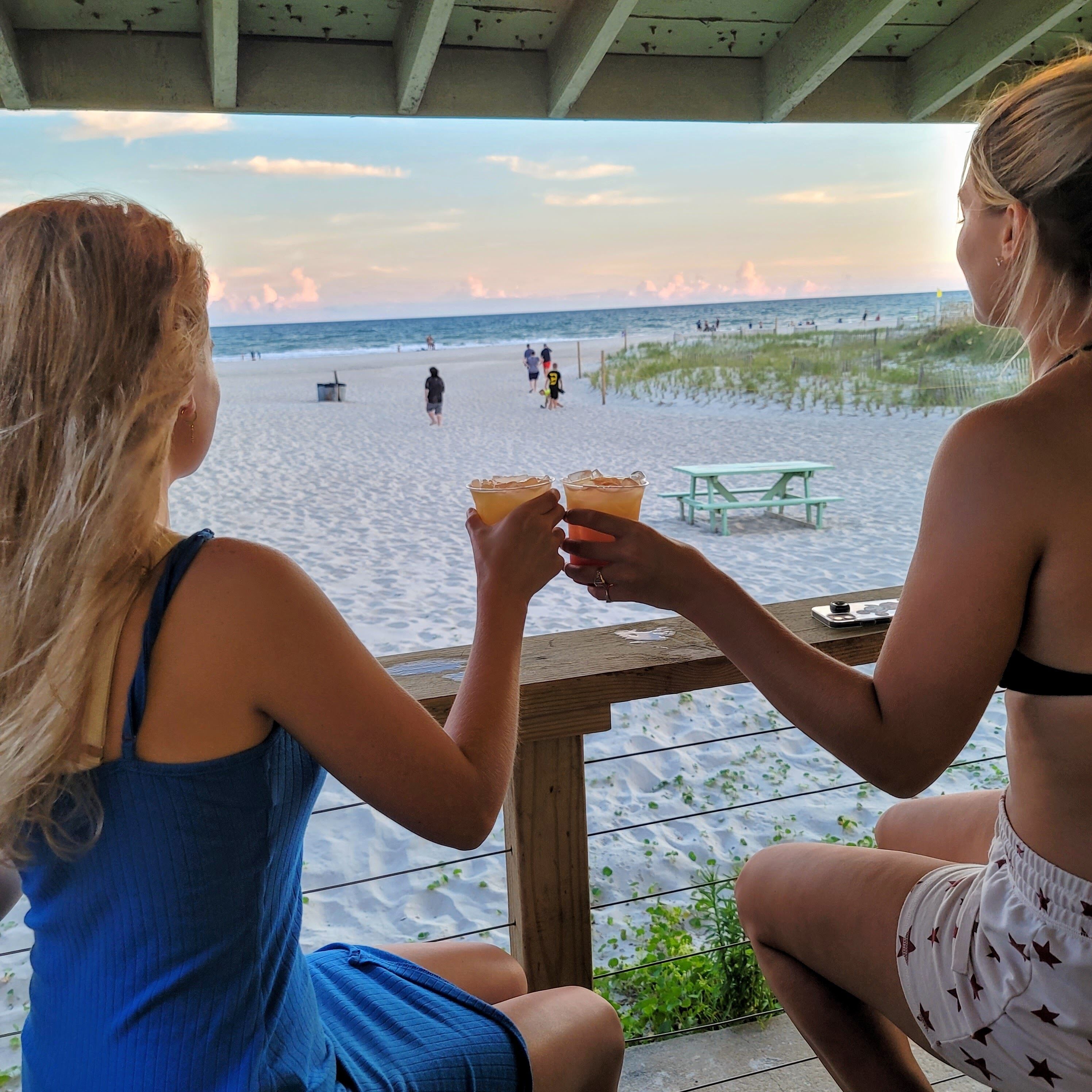 Two individuals seated at a wooden table overlooking a beach with a view of the ocean and people in the distance.