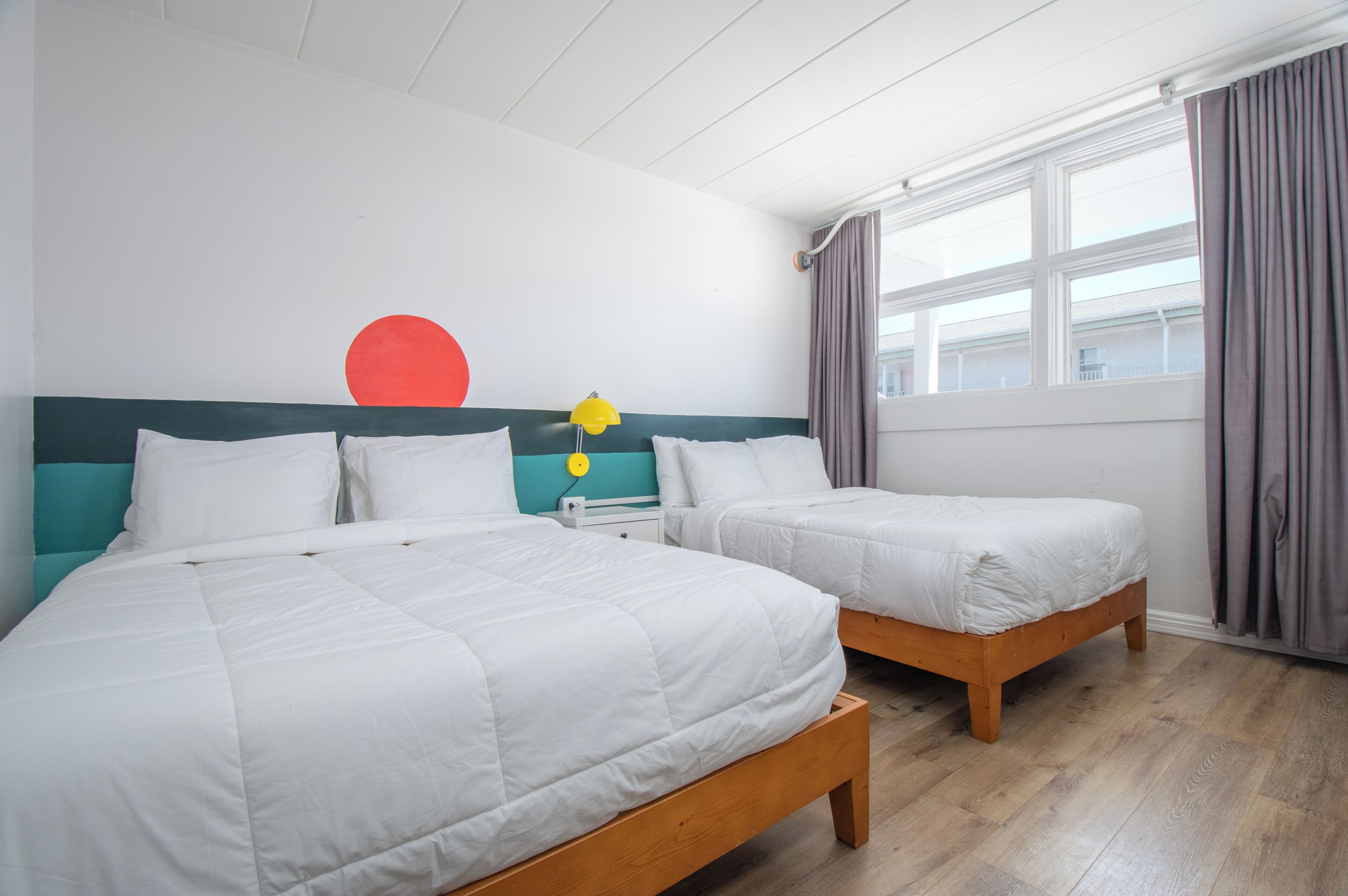 A hotel room with two beds, a window with curtains, a wall with a red circle and a yellow star, and a wooden floor.