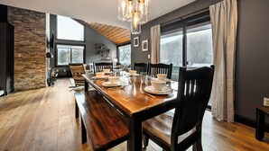 Een eetruimte met een houten tafel voor vier personen, omringd door stoelen, een stenen muur en een groot raam met gordijnen.